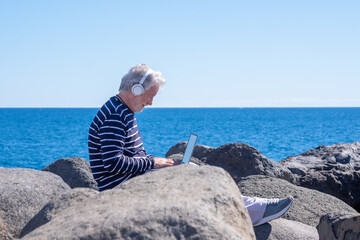 Senior caucasian man sitting on the rocks at sea using laptop in remote working, white-haired elderly male browsing with computer