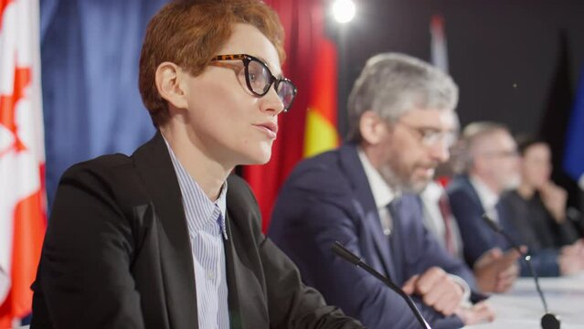 Female diplomat in glasses and formal outfit sitting at press conference table with other participants and talking to journalists