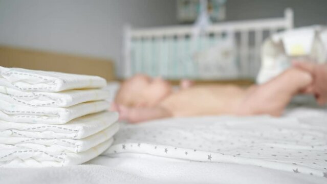 Mother hands carefully dress baby girl lying on bed in new diaper at home on blurred background. Stack of clean diapers on foreground selective focus