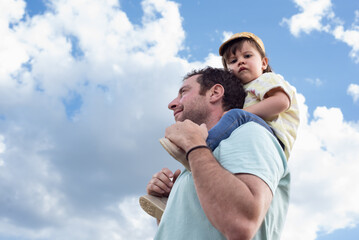 Low angle view of a little girl sitting on her father's shoulders looking at camera with a serious gesture on a background of blue sky with clouds. 