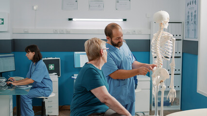 Physiotherapist pointing at spinal cord on human skeleton, explaining physical diagnosis to senior...