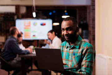 Portrait of smiling african american office worker holding laptop in business office during late night meeting working overtime. Startup employee posing confident in busy workspace with mixed team