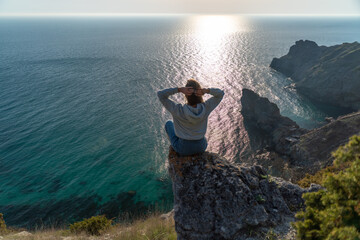 Obraz premium Woman tourist enjoying the sunset over the sea mountain landscape. Sits outdoors on a rock above the sea. She is wearing jeans and a blue hoodie. Healthy lifestyle, harmony and meditation