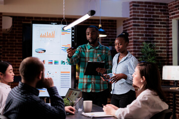 African american work colleagues brainstorming marketing strategy with sales team working overtime in late night meeting. Two coworkers discussing business ideas with board members in startup office.