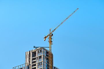 Top of the yellow tower crane on the construction of white with glass skyscraper against a blue sky with smaller crane at the bottom in sunny weather