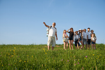 Front panoramic view of old man showing nature to group of kids. Male with gray hair and beard telling, pointing landscape by hand. Concept of active recreation and hiking.