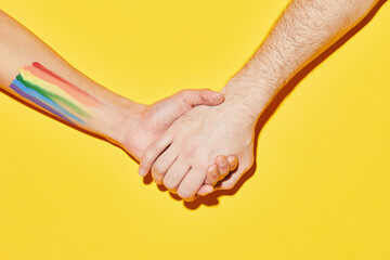 Vibrant shot of two people holding hands with LGBT flag tattoo on yellow background