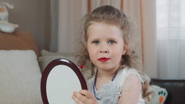 Cheerful little girl paints her lips with bright red lipstick borrowed from her mother and make kiss to camera. Funny baby does her own makeup, looking merrily in the mirrora. High quality 4k footage