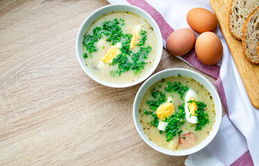 Zurek is a traditional Polish soup. Two bowls of hot soup with egg and sausage, bread and fresh herbs on the table.
