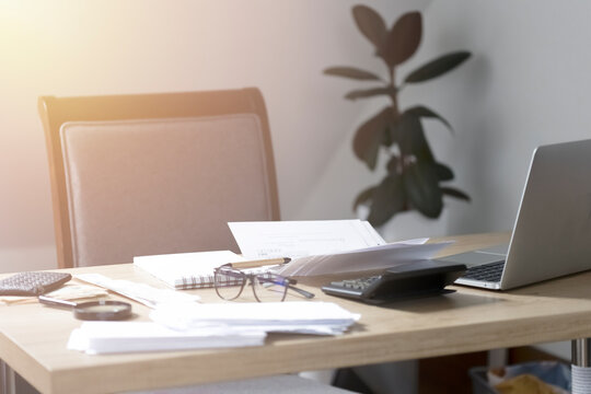 Office of an accountant or businessman. On the desktop are papers, a calculator, a laptop, glasses and a notepad. Sunlight shines through the window onto a desktop with papers and ficus in the backgro