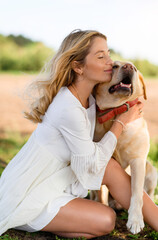 Portrait of a young beautiful blonde girl kissing her light colored Labrador dog at sunset in a field