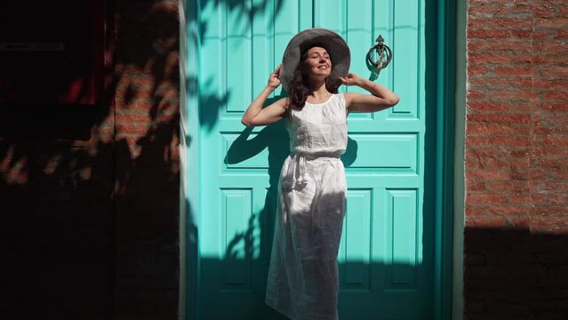 Wide shot carefree beautiful woman in white dress standing at green door and red wall in ancient town looking away. Portrait of happy Caucasian tourist enjoying summer leisure on Cyprus outdoors