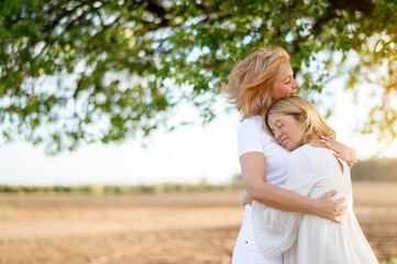 Portrait of a beautiful blonde mother with her blonde daughter hugging in a field at sunset where the wind blows on them