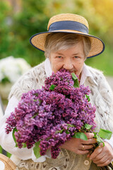 Beautiful cute old grandmother with a hat with a bouquet of lilacs in her hands looks and smiles on the background of the garden