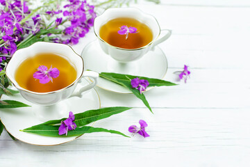 Two cups of infusion (drink, decoction) of ivan-tea on a white wooden background. Grass and flowers of the plant kiprey, ivan-grass, epilobium. Space for the text.