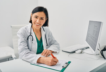 Portrait of general practitioner woman wearing doctor's coat sitting in her workplace office at medical clinic. Doctor's consultation