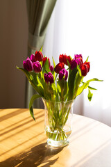 Glass vase on the table with tulips, light from the window and shadows. Place for text, romantic. The concept of birthday, holiday, romance, good morning, mother's day, March 8, valentine's day,