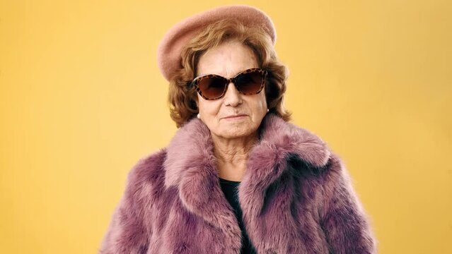 Stylish senior woman with a fur coat and sunglasses smiling while standing over an yellow background.