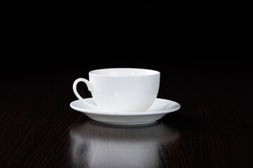 A clean, white, empty cup and saucer stands on a table against a black background.