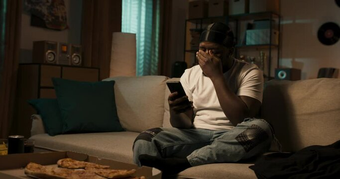 Relaxed dark skinned man in white T-shirt and black bandana relaxes on couch with cell phone in hand, bearded guy is sad argues over text messages.