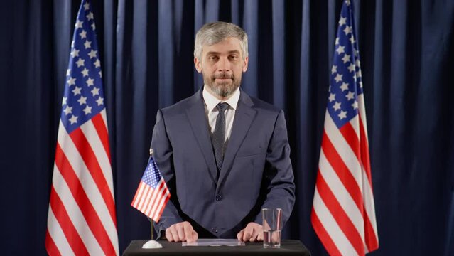 Middle aged male politician with gray hair and beard looking at camera and giving speech at rostrum with US flag on it during press conference