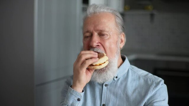 Close-up Portrait Of Disgruntled Mature Adult Male Eating Unappetizing Burger At Kitchen Home. Hungry Older Bearded Disappointed With Taste Of Bad Quality Cheeseburger. Shooting In Slow Motion.