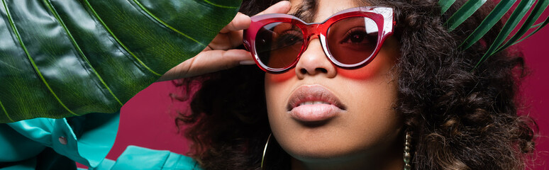 african american woman with wavy hair adjusting stylish sunglasses near leaves isolated on pink, banner.