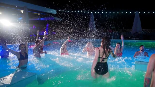 Friends have night pool party in a private villa swimming pool. Cheerful young people in swimwear splashing water, dancing and partying in luxury resort. Happy men and women hanging out. Slow motion.