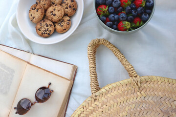Picninc blanket with straw bag, bowl of strawberries and blueberries, bowl of chocolate chip cookies, books, sunglasses and basket of apples in the garden. Top view.