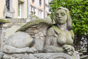 Fototapeta na wymiar Sphinx sculpture, a fusion of a lion and a woman made from stone on the street of Old Town in Gdansk, Poland. Mysterious woman sphinx statue lies near old building in the park