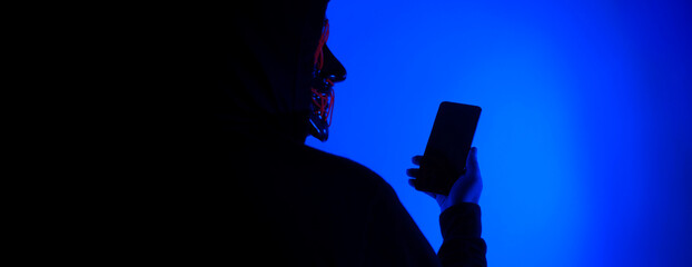 Anonymous hacker and face mask with smartphone in hand. Man in black hood shirt trying to hack...