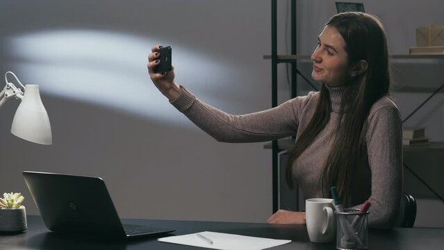 Phone selfie. Work break. Gadget leisure. Happy woman worker posing taking photo on smartphone camera at office desk workplace at evening.
