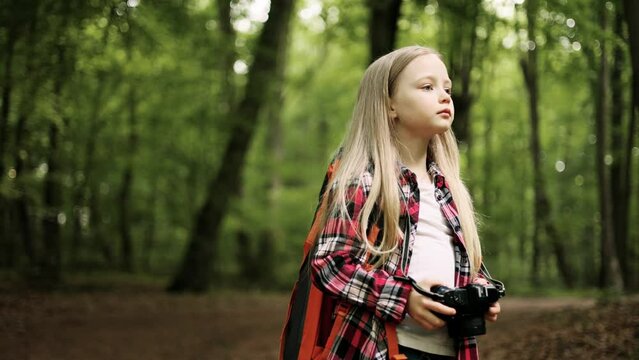 Little girl taking photo by professional digital camera in the forest. Child with a backpack explores wildlife, a kid stands alone among the trees.
