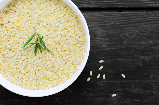 Raw risoni pasta in a white bowl with a sprig of rosemary on a wooden table. The concept of Italian cuisine. Risoni is suitable for salads and soups. Horizontal orientation. Top view. copy space.