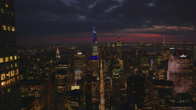Backwards fly above night city. Illuminated tall downtown skyscrapers against colourful twilight sky. Manhattan, New York City, USA