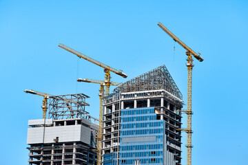 Construction of two modern skyscrapers with windows on one of them with yellow tower cranes along the buildings in bright sunny weather against a clear blue sky