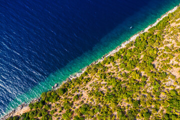Coastal area with blue clear water and forest on land, aerial view taken by drone. Half land half sea on a diagonal line. A picturesque place where transparent turquoise water meets a stony shore.