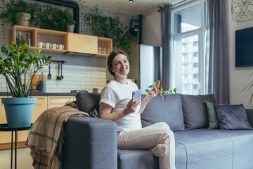 Maternity leave. Pregnant woman sitting on the couch at home, holding a mobile phone and credit card, in home clothes, ordering baby products online, choosing, buying clothes and toys for the baby.