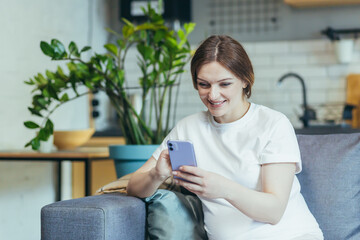 Maternity leave. Pregnant beautiful woman sitting on the couch at home, holding a mobile phone, corresponding in parent chats, typing messages, calling, watching, receiving good news. Smiling, happy