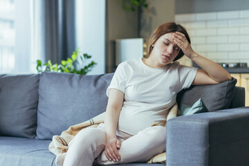 Obraz premium Feeling unwell. A pregnant woman is ill, has a headache, holds her hand behind her head. Sitting at home in home clothes on the couch
