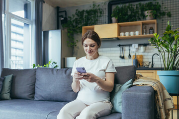 Maternity leave. Pregnant beautiful woman sitting on the couch at home, holding a mobile phone, corresponding in parent chats, typing messages, calling, watching, receiving good news. Smiling, happy
