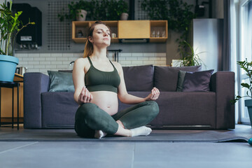 Pregnant beautiful woman doing yoga at home, sitting on a sports mat in the lotus position, closing her eyes, meditating.