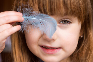 Portrait of a blurred little girl close-up and a feather of a bird in her hands stroking her cheek, gentle touch of smooth soft skin, selective focus