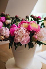 Big fresh pink and white peony blooming bouquet in a white metal bucket on a wooden table in a light bright modern beautiful apartment. Spring home decor or present idea. Vivid summer colors.