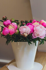 Big fresh pink and white peony blooming bouquet in a white metal bucket on a wooden table in a light bright modern beautiful apartment. Spring home decor or present idea. Vivid summer colors.