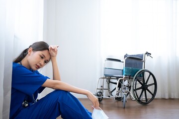 Tired exhausted female asian scrub nurse wears face mask blue uniform sits on hospital floor....