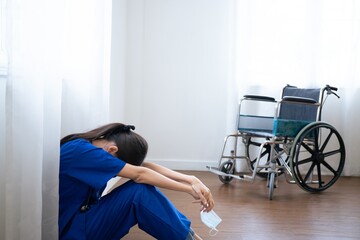 Tired exhausted female asian scrub nurse wears face mask blue uniform sits on hospital floor. Depressed sad Thai ethic doctor feels fatigue burnout stress, lack of sleep, napping at work. - obrazy, fototapety, plakaty