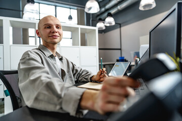 Naklejka na ściany i meble Young bald business man sitting at desk in office, working on computer