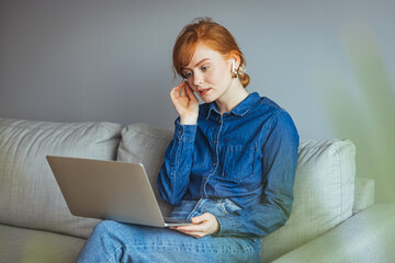 Young female resting on sofa holds on lap laptop looking at screen writing e-mail, concept of e-commerce usage, websites surfing, doing remote work at home seated on comfy couch in living room..