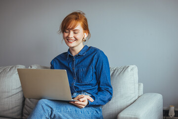 Young female resting on sofa holds on lap laptop looking at screen writing e-mail, concept of e-commerce usage, websites surfing, doing remote work at home seated on comfy couch in living room..